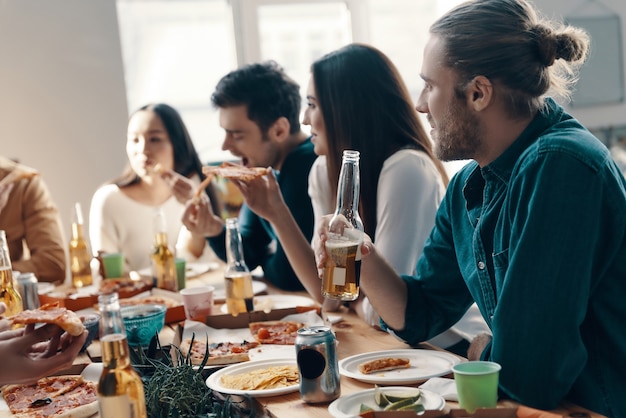 Ótimos momentos com os amigos. Grupo de jovens em trajes casuais comendo pizza e sorrindo durante um jantar dentro de casa