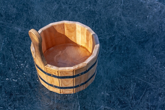 Una tina de madera con hielo en el interior está parada sobre el hielo con grietas. Los anillos de ajuste de hierro conectan partes de la bañera. Soleado. Horizontal.