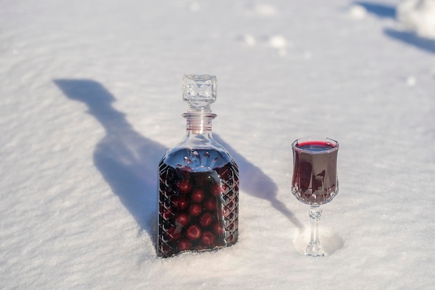 Tintura caseira de cereja vermelha em uma garrafa de vidro e uma taça de cristal de vinho sobre neve e fundo branco