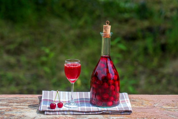 Foto tintura casera de cereza roja concepto de bebidas alcohólicas de bayas vino rojo casero hecho de cerezas maduras en una botella de vidrio y un vaso de vino en una mesa de madera en un jardín verde de fondo ucrania