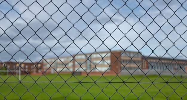 Un típico edificio de la escuela secundaria y campo de deportes bajo un cielo nublado con foco en la valla de seguridad en primer plano