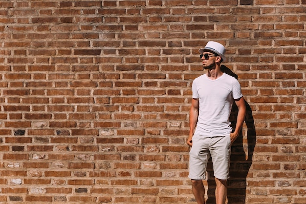 Un tipo con barba vestido de blanco y gafas de sol se toma de la mano en los bolsillos cerca de la pared de ladrillo