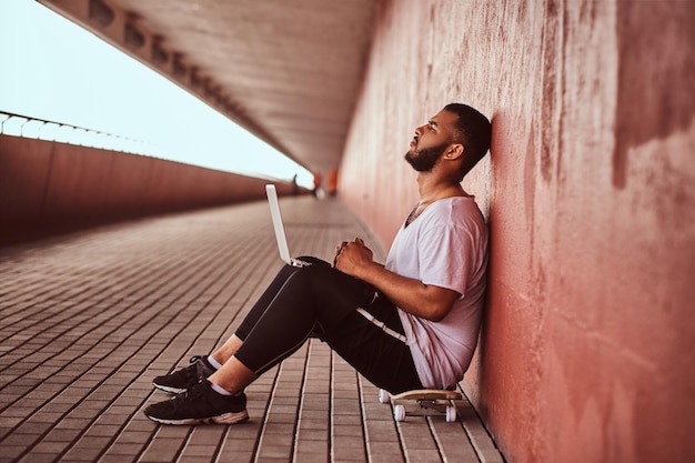 Un tipo barbudo afroamericano pensativo vestido con una camisa blanca y pantalones cortos deportivos sostiene una laptop mientras se sienta en una patineta debajo de un puente, apoyado en una pared.