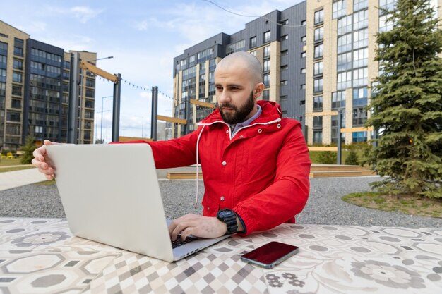 Un tipo brutal con barba y chaqueta roja está trabajando en una computadora en la calle. Concepto de empleo online