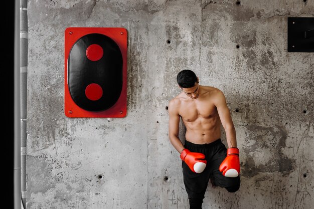 Un tipo brutal de cabello oscuro con el torso desnudo y los guantes de boxeo rojos está parado contra una pared de concreto junto a un equipo de boxeo.