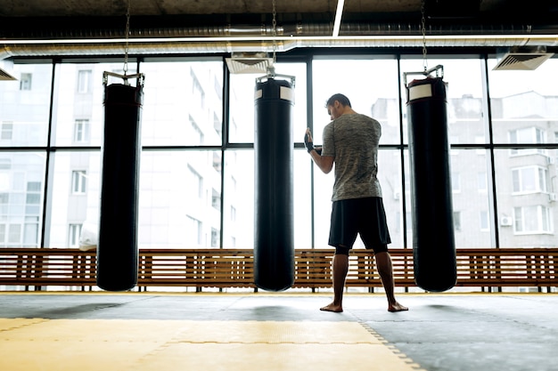 Foto un tipo de cabello oscuro vestido con la camiseta gris y pantalones cortos negros se encuentra junto a los sacos de boxeo en el contexto de las ventanas panorámicas en el gimnasio de boxeo.