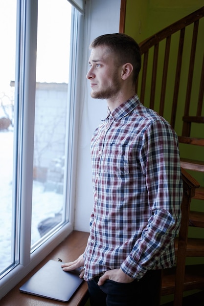 Un tipo con una camisa a cuadros con una computadora portátil mira por la ventana