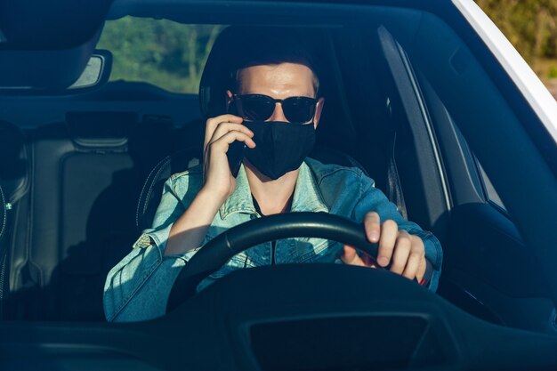 Un tipo con una chaqueta de mezclilla y gafas negras con una máscara en la cara está hablando por teléfono mientras conduce un automóvil