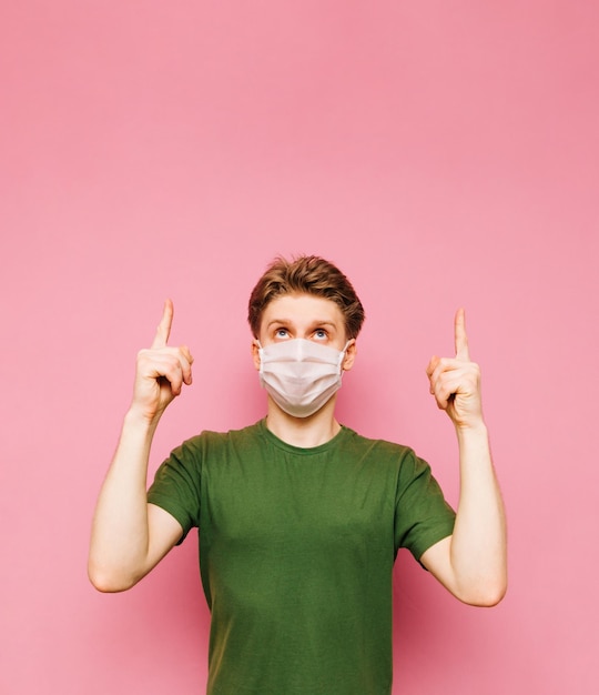 Un tipo emocional con camiseta verde y una máscara médica en la cara mira y señala con los dedos hacia arriba