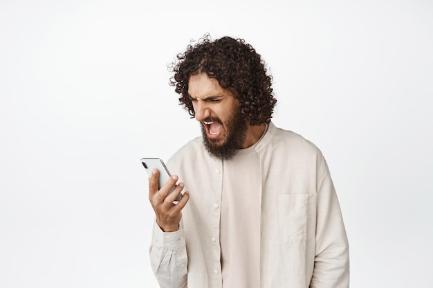 Un tipo enojado de Oriente Medio gritando a su teléfono móvil luciendo enojado con el fondo blanco del teléfono inteligente