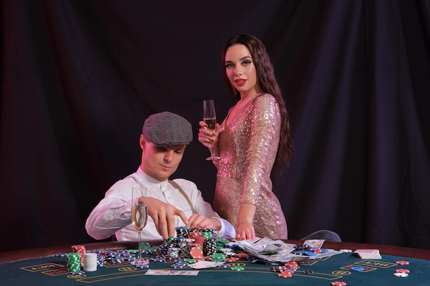 El tipo está jugando al póquer en el casino sentado en la mesa con montones de fichas de tarjetas de dinero Celebrando la victoria con una mujer joven y hermosa Retroiluminación de colores de fondo negro Apuestas de alcohol Primer plano