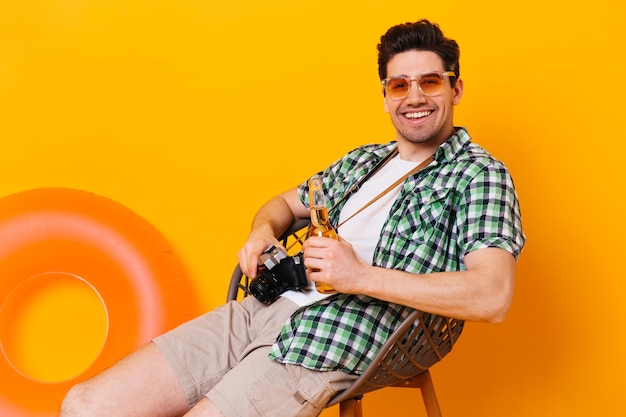 Un tipo feliz con gafas naranjas se relaja en una silla de madera sonriendo sosteniendo una botella de cerveza y una cámara retro