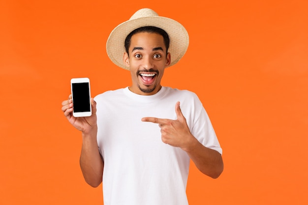 Foto un tipo feliz y sonriente que muestra la aplicación de la agencia de viajes, el hotel reservado a precios excelentes, muestra la pantalla del teléfono y señala la aplicación para promocionar, compró un viaje de vacaciones, pared naranja