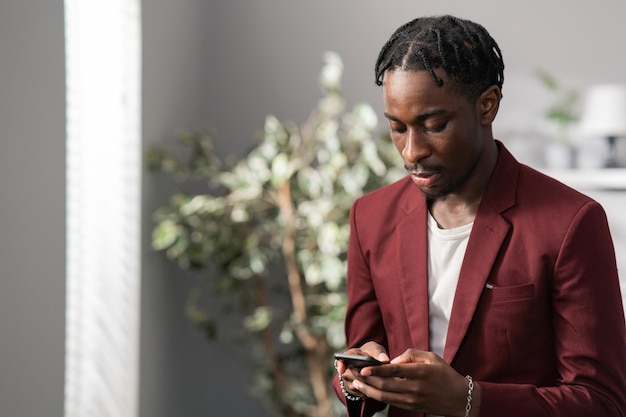 Un tipo guapo con rastas está parado junto a la ventana de la oficina con una chaqueta granate