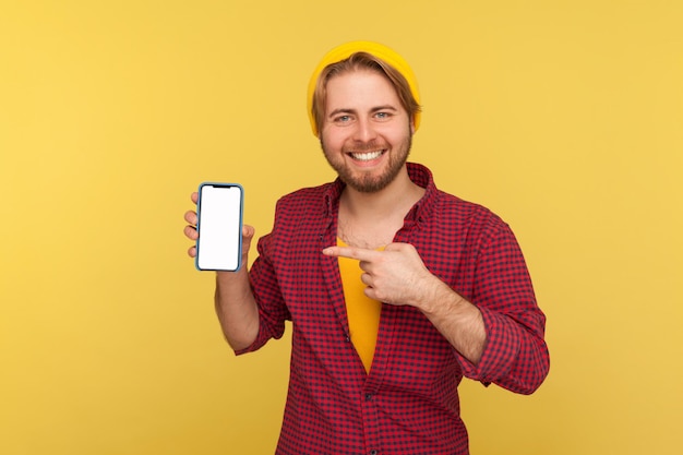 Un tipo hipster de moda feliz con sombrero de beanie y camisa a cuadros apuntando a la pantalla vacía del teléfono móvil, mirando a la cámara con una sonrisa dentuda, mostrando una maqueta en blanco del teléfono móvil para publicidad. tiro del estudio aislado