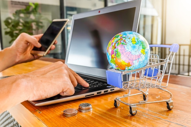 Foto tipo de mano recortada en una computadora portátil por globo en un carrito de compras en miniatura y usando un teléfono inteligente sobre la mesa