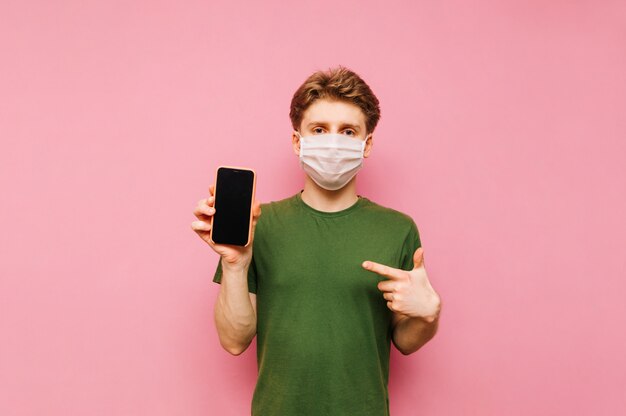 Un tipo con una máscara de gasa se para con un teléfono inteligente en la mano en una rosa, señala con el dedo a la pantalla negra y mira a la cámara con una cara seria