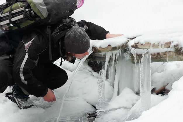 Un tipo con mochila bebe agua fría de una fuente. Caminata de invierno