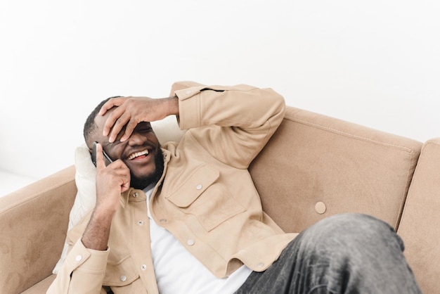 Un tipo negro emocionado está usando un teléfono inteligente hablando con un amigo mientras está acostado en el sofá en casa Conexión móvil