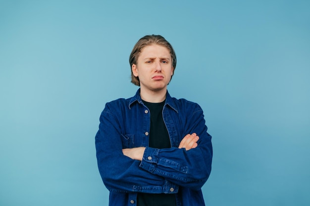 Un tipo ofendido con cabello largo y una camisa azul se para contra la pared azul y se ve infeliz