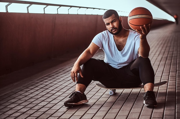 Un tipo pensativo de piel oscura vestido con una camisa blanca y pantalones cortos deportivos sostiene una pelota de baloncesto mientras se sienta en una patineta en una acera debajo de un puente, mirando una cámara.