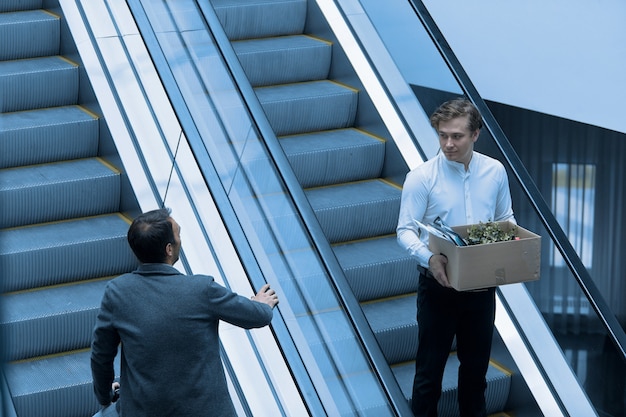 Un tipo que es despedido del trabajo baja por las escaleras mecánicas con una caja de cosas más allá de su jefe.