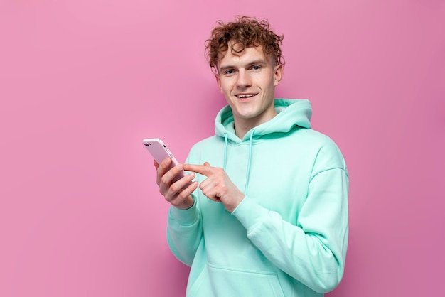 Un tipo rizado con una sudadera de menta usa un teléfono inteligente en un fondo rosado aislado un hombre con una camiseta azul
