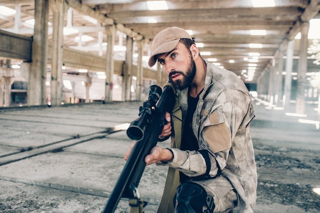 Un tipo serio está prestando atención a los sonidos en un gran edificio vacío. El esta en el hangar. El soldado sostiene un gran rifle y mira hacia adelante. El hombre está listo para disparar.