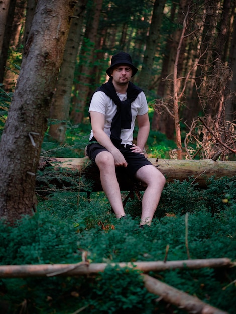 Un tipo con un sombrero negro y una camiseta blanca se sienta en un tronco en el bosque y sonríe