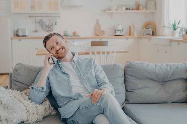 Un tipo sonriente se sienta en el sofá de casa divirtiéndose mientras habla por un teléfono móvil moderno