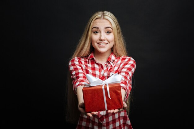 Tipo, um adolescente sincero e encantado expressando felicidade e dando uma caixa de presente enquanto está isolado na parede preta