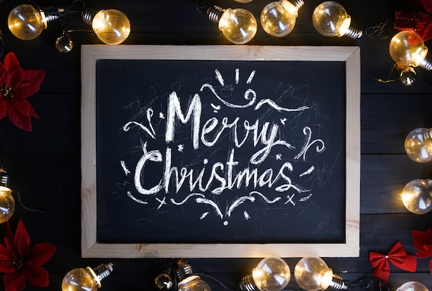 Tipografia de feliz Natal no quadro-negro entre lâmpadas e poinsettia vermelho em madeira preta