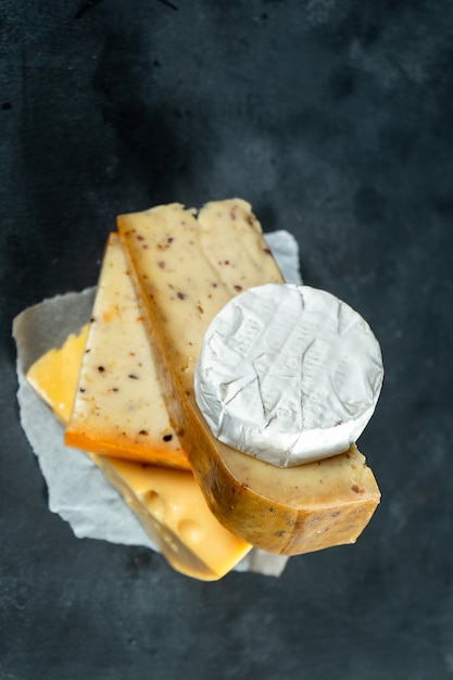 Tipos diferentes criativos de queijo que colocam no fundo escuro com espaço da cópia. Camembert, queijo com especiarias, queijo holandês. Grande cartaz para loja de queijos. Fundo de alimentos. Foco suave