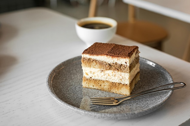 Tiramisu-Dessertkuchen auf einem Teller mit einer Gabel und einer weißen Tasse schwarzen Kaffee am Tisch eines Cafés Lifestyle-Bild, selektiver Fokus, geringe Schärfentiefe, Bokeh-Hintergrund