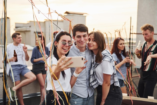 Tirando selfie grupo de jovens em roupas casuais fazem uma festa no telhado juntos durante o dia