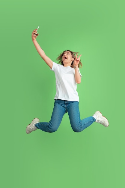 Tirando selfie no salto. Retrato de mulher jovem branca isolado na parede verde. Bela modelo feminino em camisa branca. Conceito de emoções humanas, expressão facial, juventude.