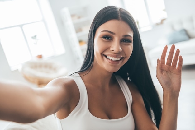 Tirando uma bela selfie. Auto-retrato de uma jovem mulher bonita olhando para a câmera e sorrindo enquanto está sentado no sofá em casa