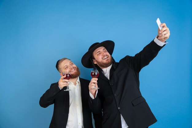 Tirando uma selfie com vinho. retrato de um jovem judeu ortodoxo isolado na parede azul. purim, negócios, festival, feriado, celebração pessach ou páscoa, judaísmo, conceito de religião.