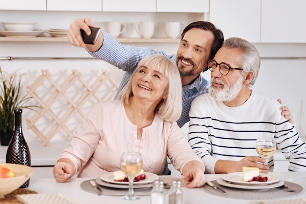 Tirando uma selfie para um álbum de família. Homem sorridente e divertido jantando e curtindo o tempo com seus pais idosos enquanto segura o gadget e faz uma selfie