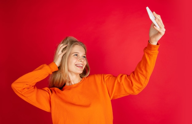 Tirando uma selfie. Retrato de uma jovem mulher caucasiana, isolada em um fundo vermelho studio com copyspace. Linda modelo feminino. Conceito de emoções humanas, expressão facial, vendas, anúncio, juventude. Folheto