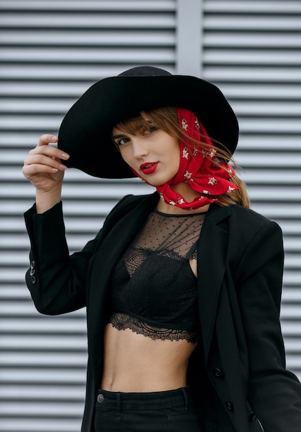 Tiro al aire libre de hermosa mujer morena con labios rojos viste traje negro, sombrero de ala ancha posando cerca de las contraventanas de metal