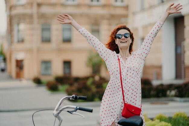 Foto tiro al aire libre horizontal de mujer pelirroja positiva encantada levanta las manos