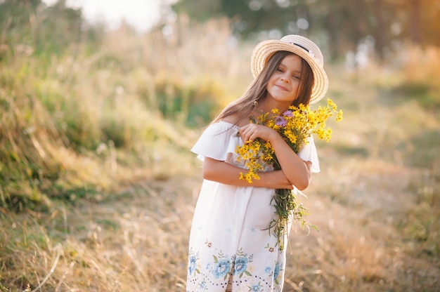 Tiro al aire libre de una joven de aspecto agradable con una piel bronceada y saludable, vestida con un vestido blanco y un sombrero de verano, posa en el parque con una expresión de confianza y satisfacción, le gusta la recreación