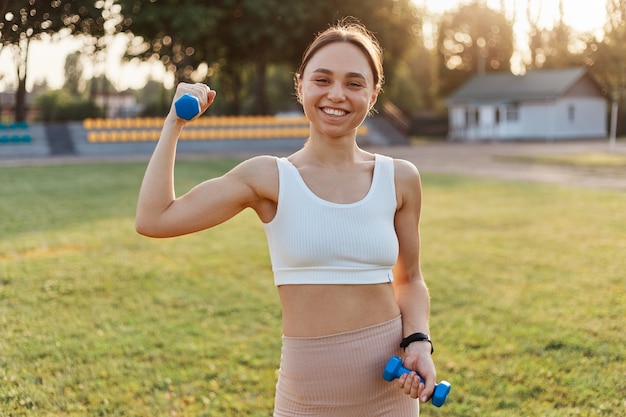 Tiro al aire libre de la joven atleta con pesas estirando y calentando en el estadio, mirando sonriendo a la cámara, expresando felicidad, deporte y fitness.