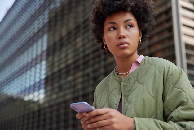 Tiro al aire libre de la modelo de mujer rizada joven pensativa utiliza el teléfono inteligente para blogs