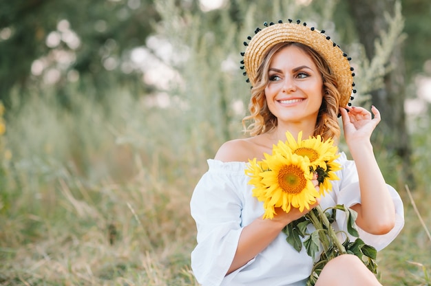 Tiro al aire libre de mujer de aspecto agradable con piel bronceada y saludable, vestida con vestido blanco y sombrero de verano, posa en el parque con expresión de confianza y satisfacción, le gusta la recreación