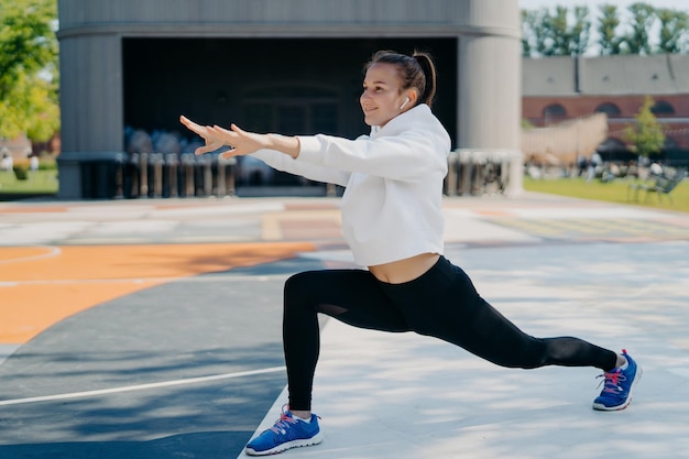 Tiro al aire libre de una mujer delgada y deportiva involucrada en actividades recreativas que estira los brazos, las piernas, mira a la distancia, usa leggings blancos con capucha y zapatos deportivos, estira el cuerpo antes de correr lleva un estilo de vida saludable