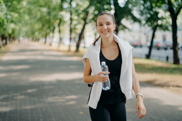 Tiro al aire libre de mujer deportiva alegre bebe agua fresca de botella tiene caminar durante el verano parque urbano verde tiene una sonrisa en la cara lleva un estilo de vida saludable. Restaurar el equilibrio acuático después del entrenamiento