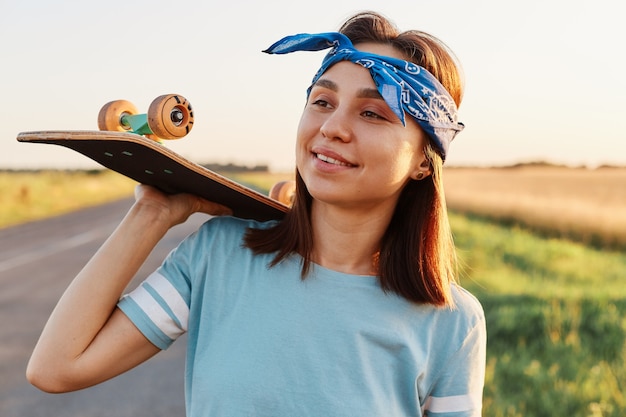 Tiro al aire libre de mujer morena sonriente con camiseta de estilo casual y banda para el cabello, sosteniendo el patín sobre los hombros, mirando a otro lado con una sonrisa, expresando felicidad.