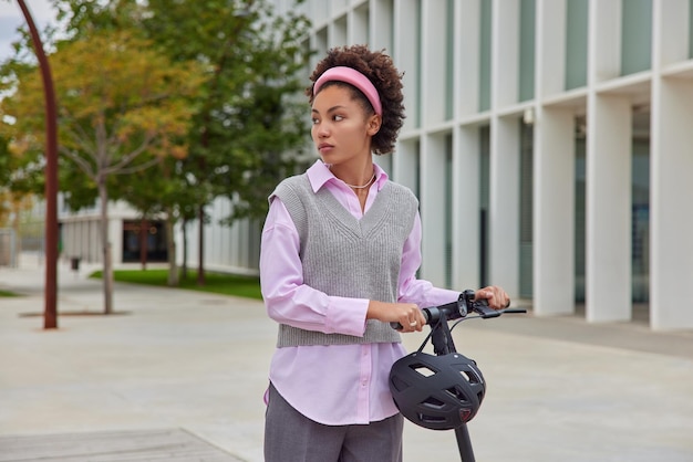 Tiro al aire libre de mujer pensativa en ropa casual paseos en scooter eléctrico en la calle pasa el tiempo libre utiliza activamente poses de casco protector en la ciudad